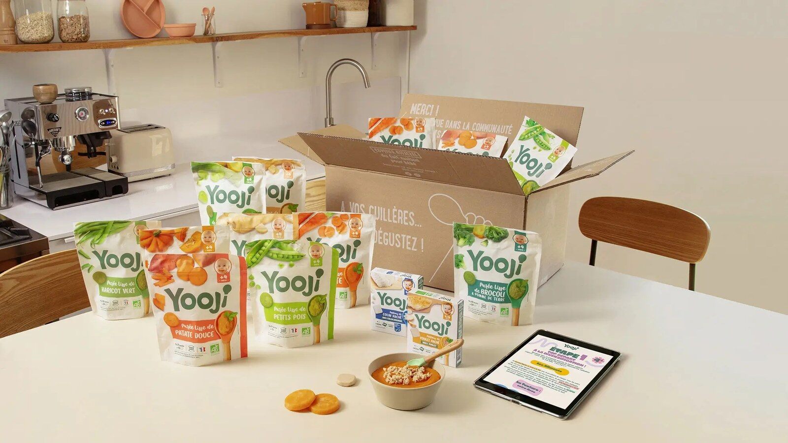 Une box Yooji ouverte sur une table de cuisine, entourée de sachets de purées lisses pour bébé, d’un bol de purée et d’une tablette