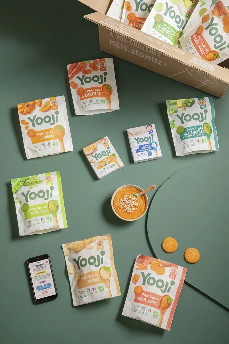 Exemple de box Yooji : sachets de purées lisses de carotte, potiron, petit pois, panais, brocoli et patate douce, étuis de protéines et un bol de purée, autour d’un carton ouvert